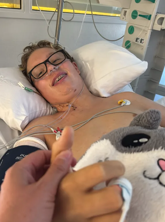 Luca liegt lächelnd in seinem Klinikbett, trägt seine Brille und eine Sauerstoff-Nasenbrille; jemand hält seine Hand, neben ihm liegt ein Kuscheltier