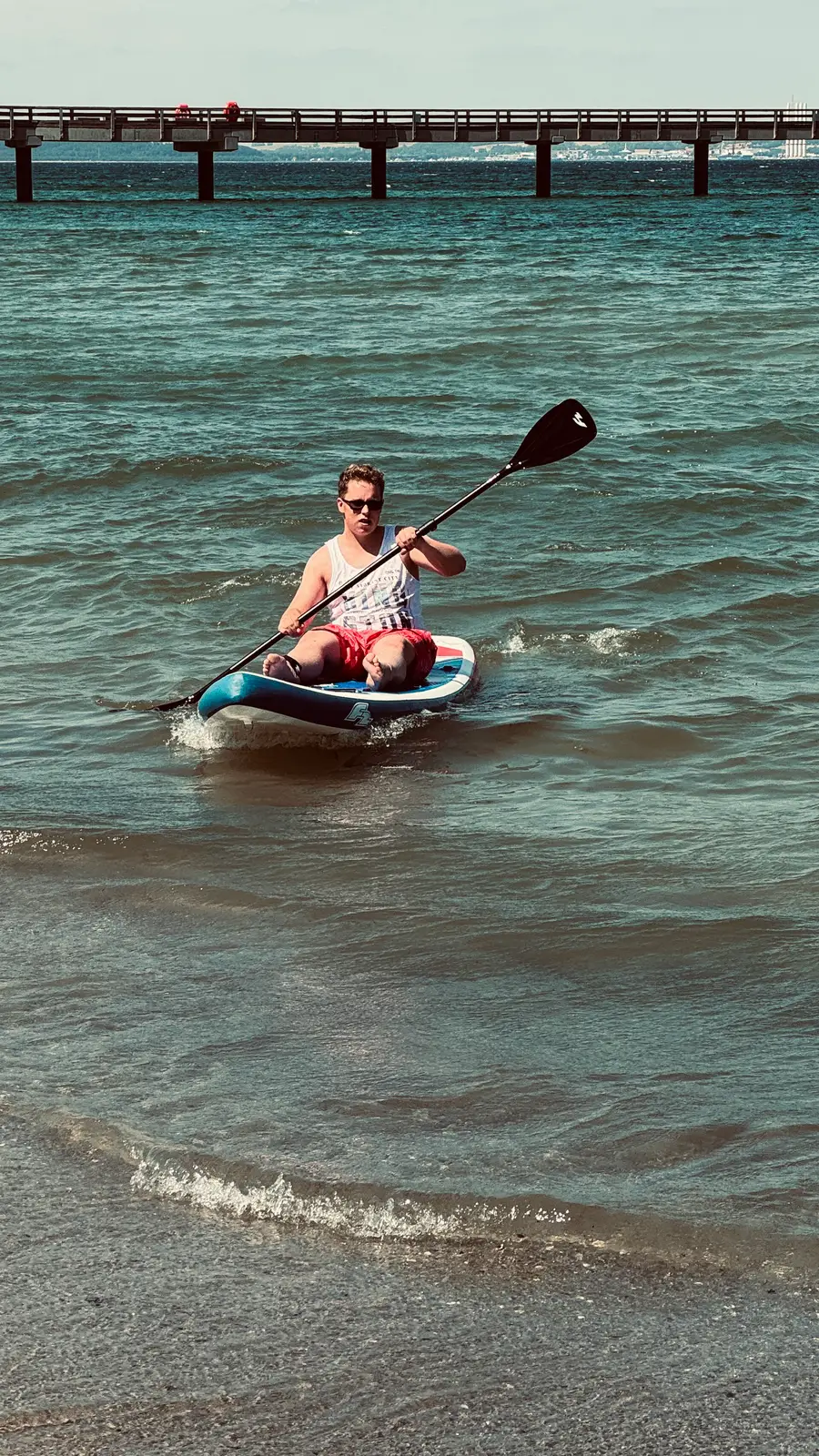 Luca paddelt kniend auf einem Stand-up-Paddle-Board vor einer Seebrücke